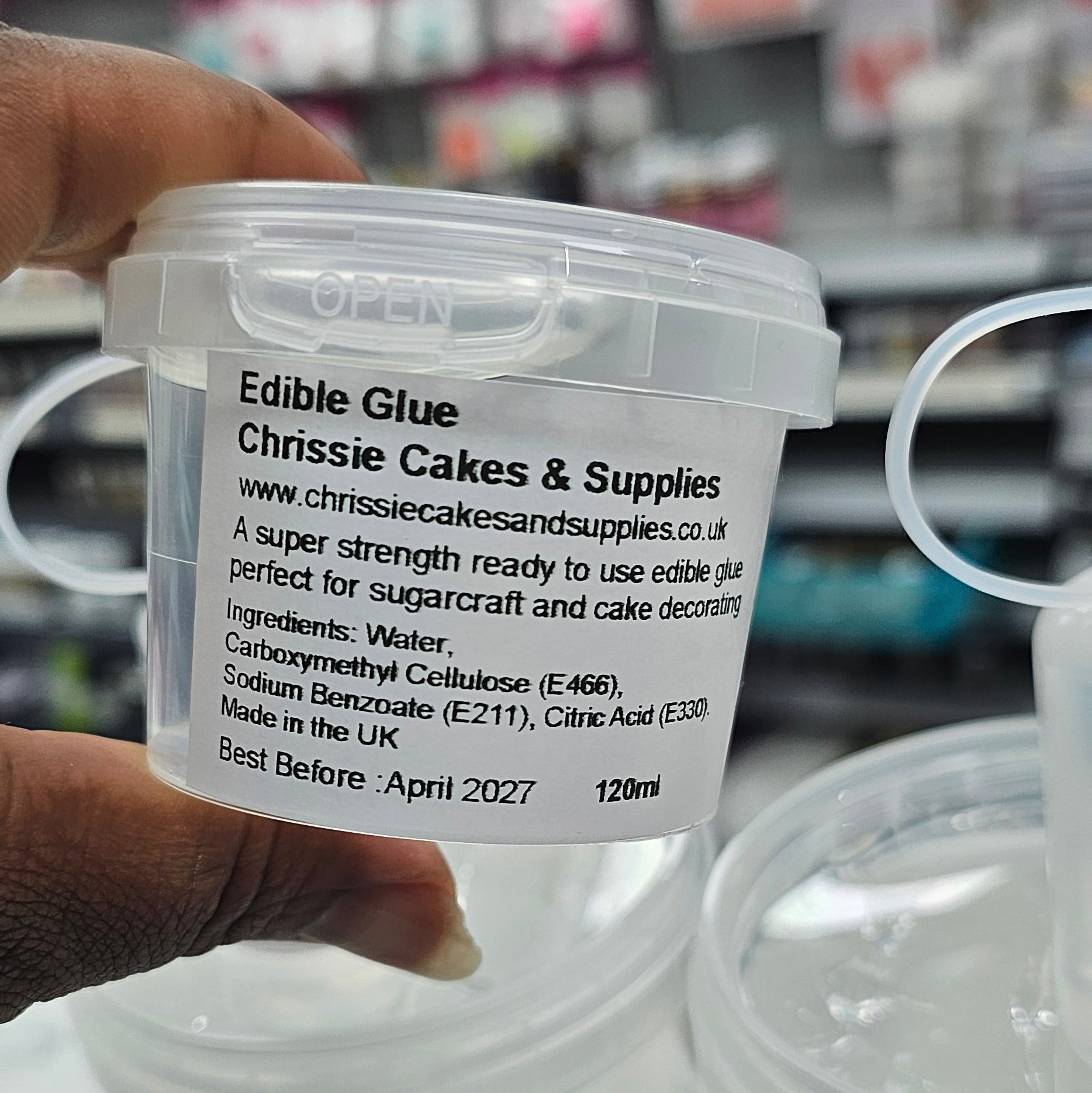 Hand holding a container of edible glue by Chrissie cakes & Supplies with a blurred store background