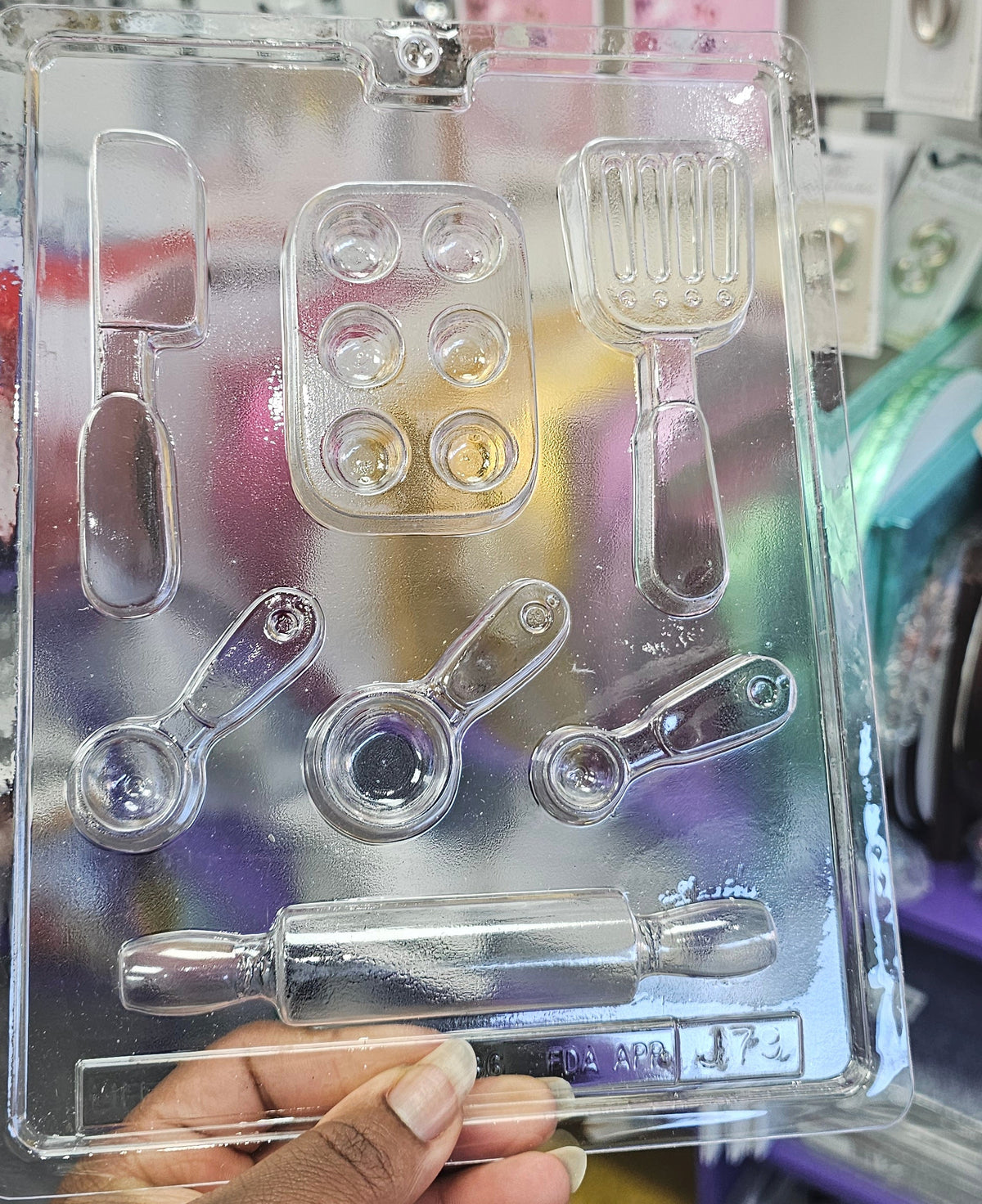A hand holding a clear plastic novelty baking utensil CHOCOLATE mould set, featuring cavities shaped like a knife, spatula, measuring cups, and a rolling pin, with a busy store shelf in the background.