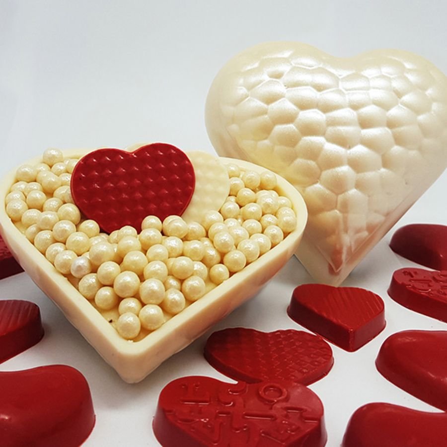 A white chocolate mould in the shape of a heart with a red textured design, displayed alongside a small red heart-shaped chocolate piece.